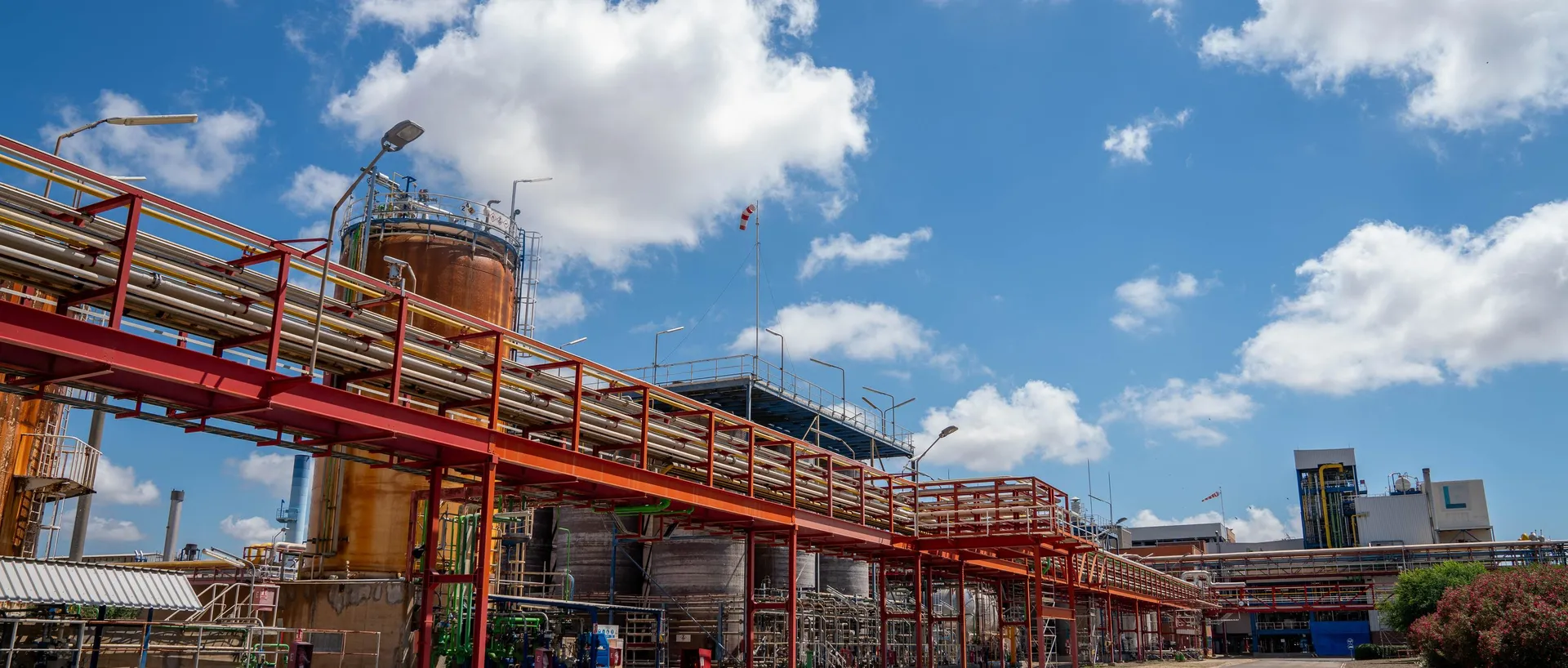 Vista de una planta petroquímica con pasarelas y tuberías metálicas elevadas de color rojo, tanques industriales y un cielo azul con nubes.