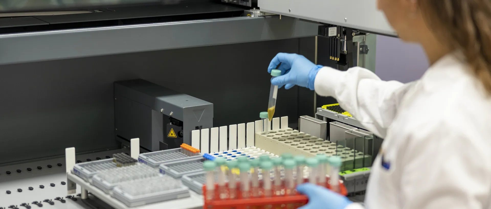Investigadora con bata blanca y guantes azules coloca un tubo con una muestra en un sistema automatizado de almacenamiento del laboratorio, con una gradilla roja de tubos en la otra mano.