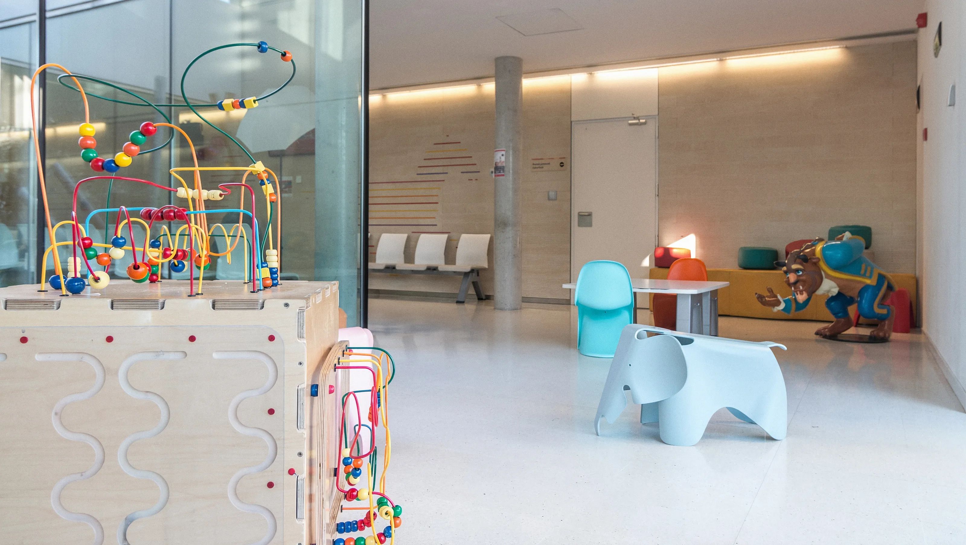 Sala de espera infantil del Hospital Sant Joan de Déu Barcelona, con un laberinto de cuentas de colores en primer plano, sillas y un taburete con forma de elefante, sin nadie en la sala.