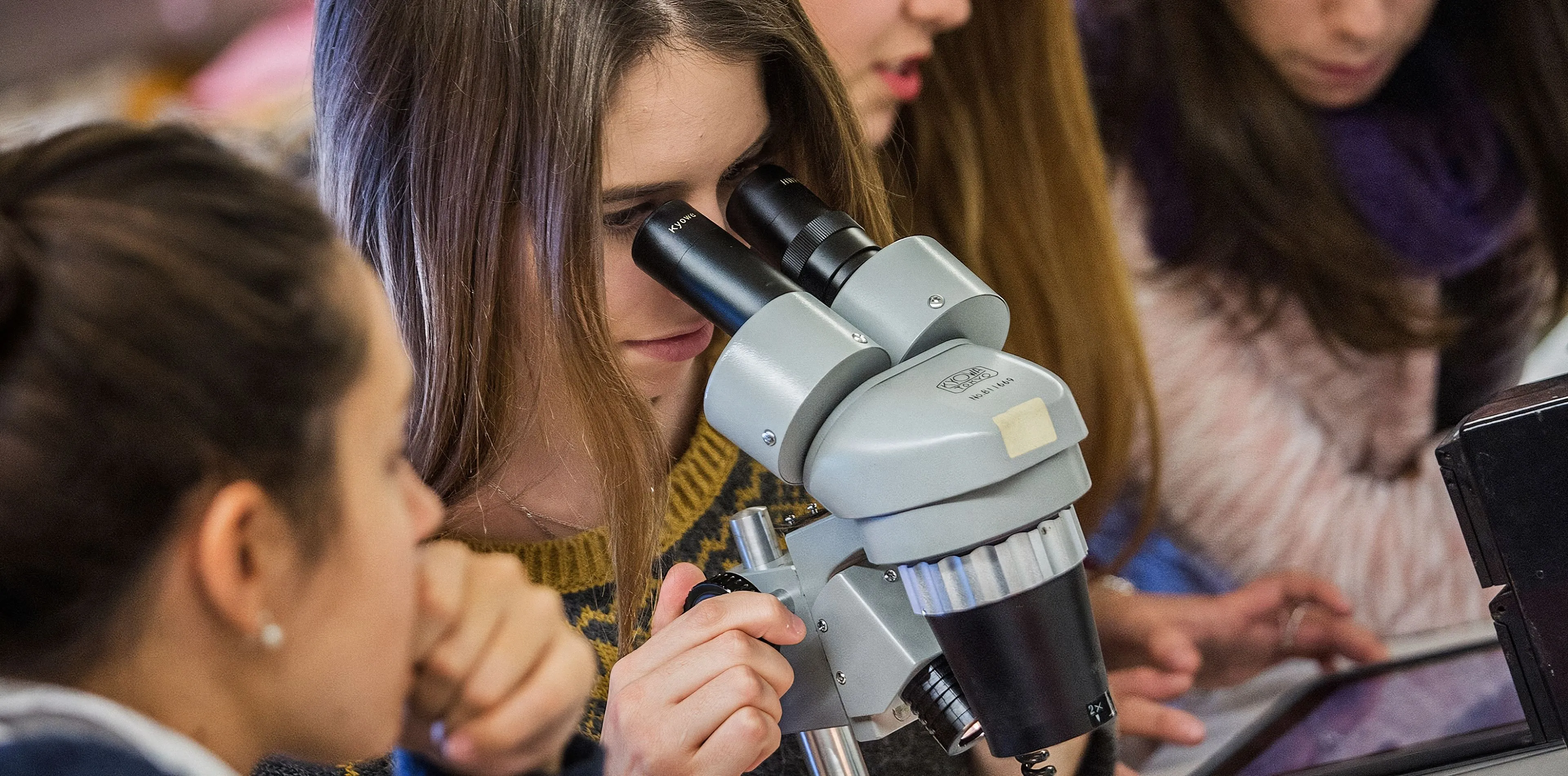 Una estudiante mira por un microscopio estereoscópico en un laboratorio, rodeada de compañeras que observan y consultan una tableta.