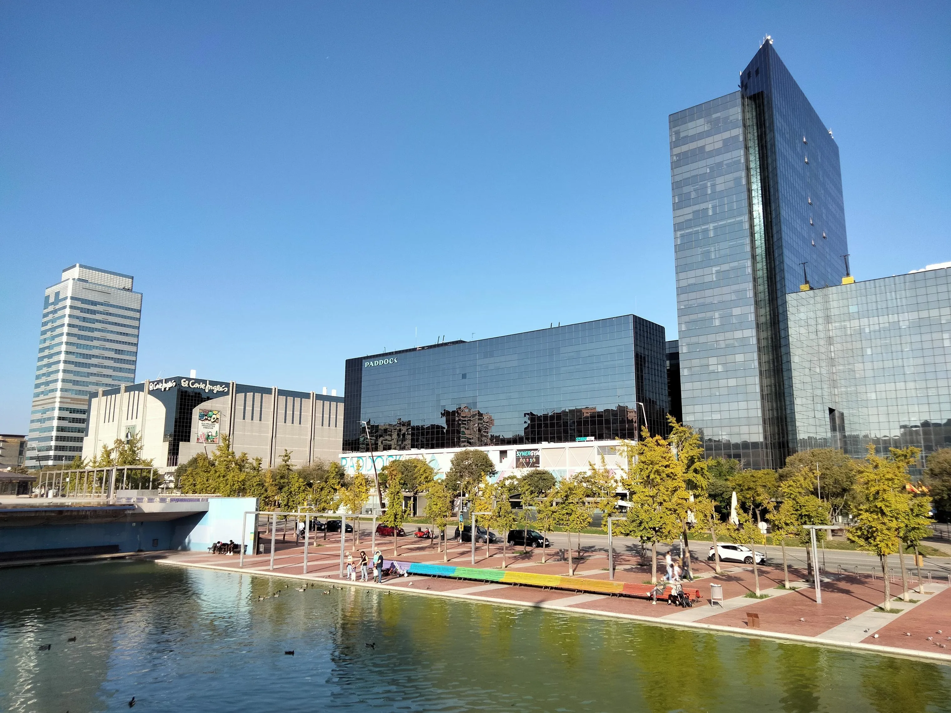 El lago del Parc de Catalunya de Sabadell con bancos de colores junto al agua y, al fondo, los rascacielos del Eix Macià y el edificio Paddock.