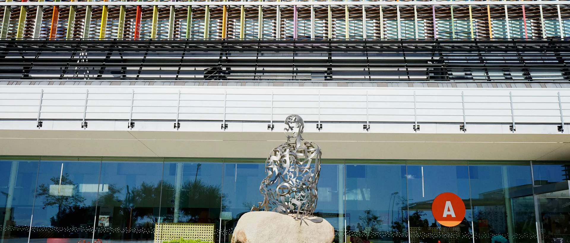 Escultura de Jaume Plensa, una figura sentada hecha de letras de acero sobre una roca, ante la fachada acristalada del Hospital Sant Joan de Déu Barcelona, con lamas de colores en las plantas superiores.