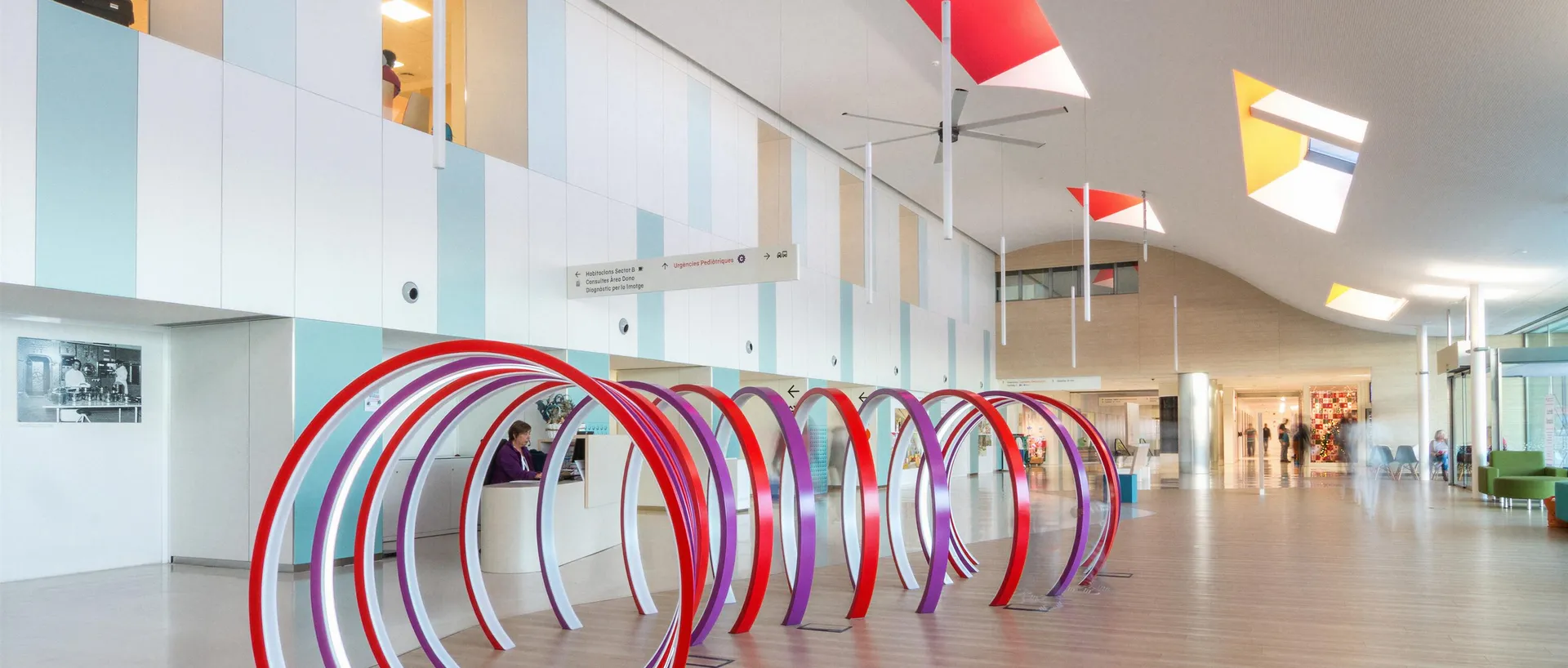 Vestíbulo principal del Hospital Sant Joan de Déu Barcelona, con una escultura de arcos rojos y morados sobre el suelo de madera, el mostrador de información al fondo y lucernarios de colores en el techo.