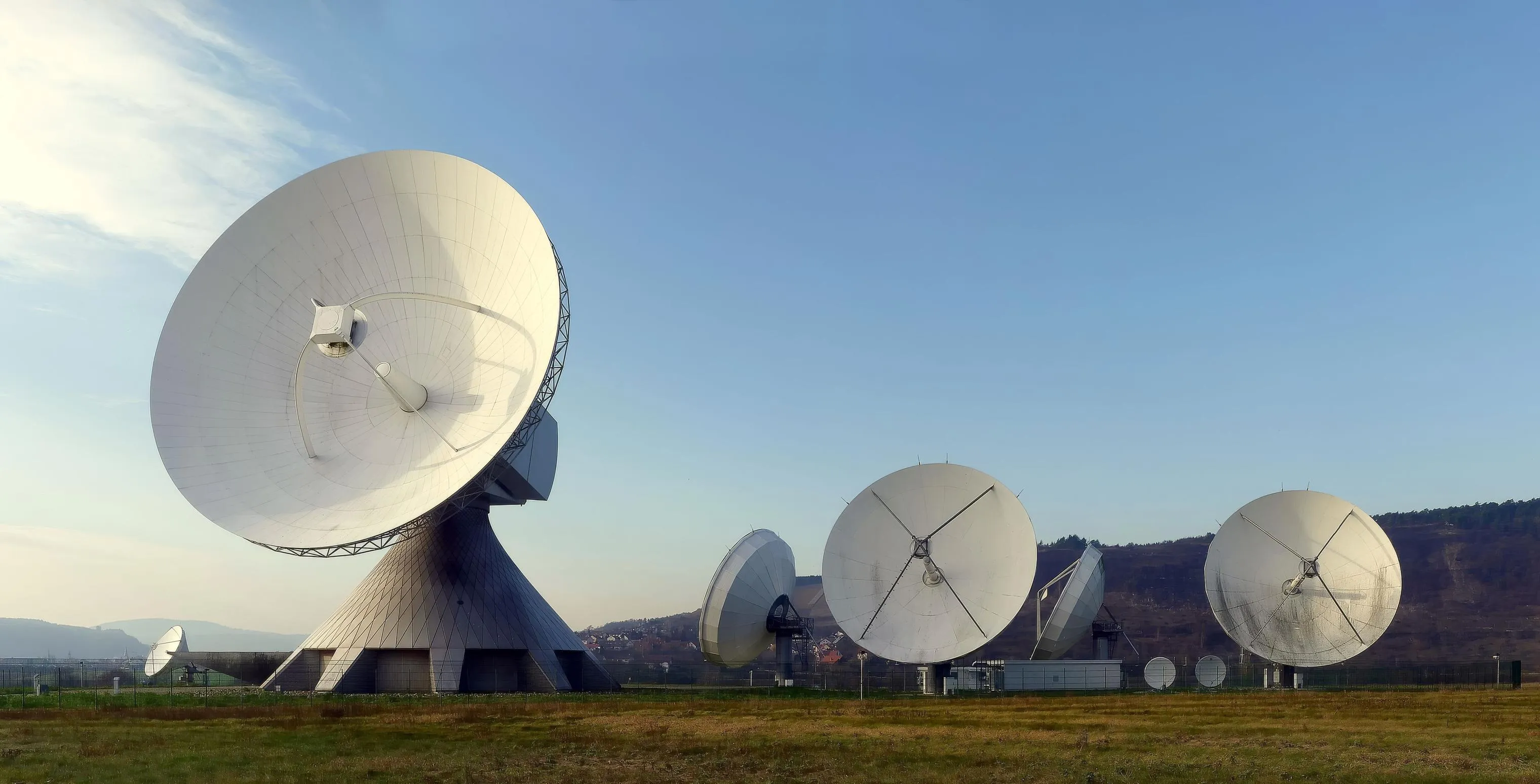 Varias antenas parabólicas blancas de gran tamaño en una estación de seguimiento por satélite, bajo un cielo despejado, sin personas.