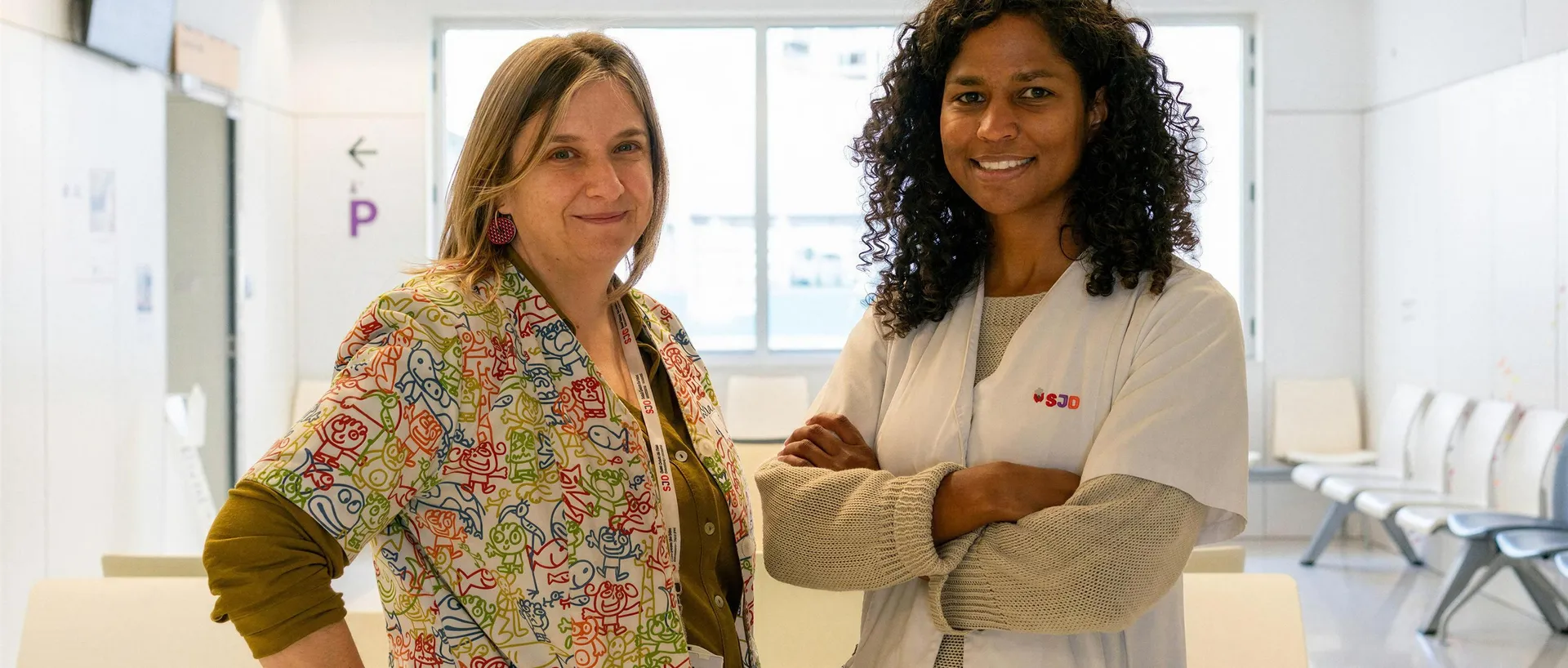 Dos profesionales del Hospital Sant Joan de Déu sonríen a cámara en una sala de espera luminosa: una con una bata estampada de dibujos infantiles y otra con bata blanca y los brazos cruzados.