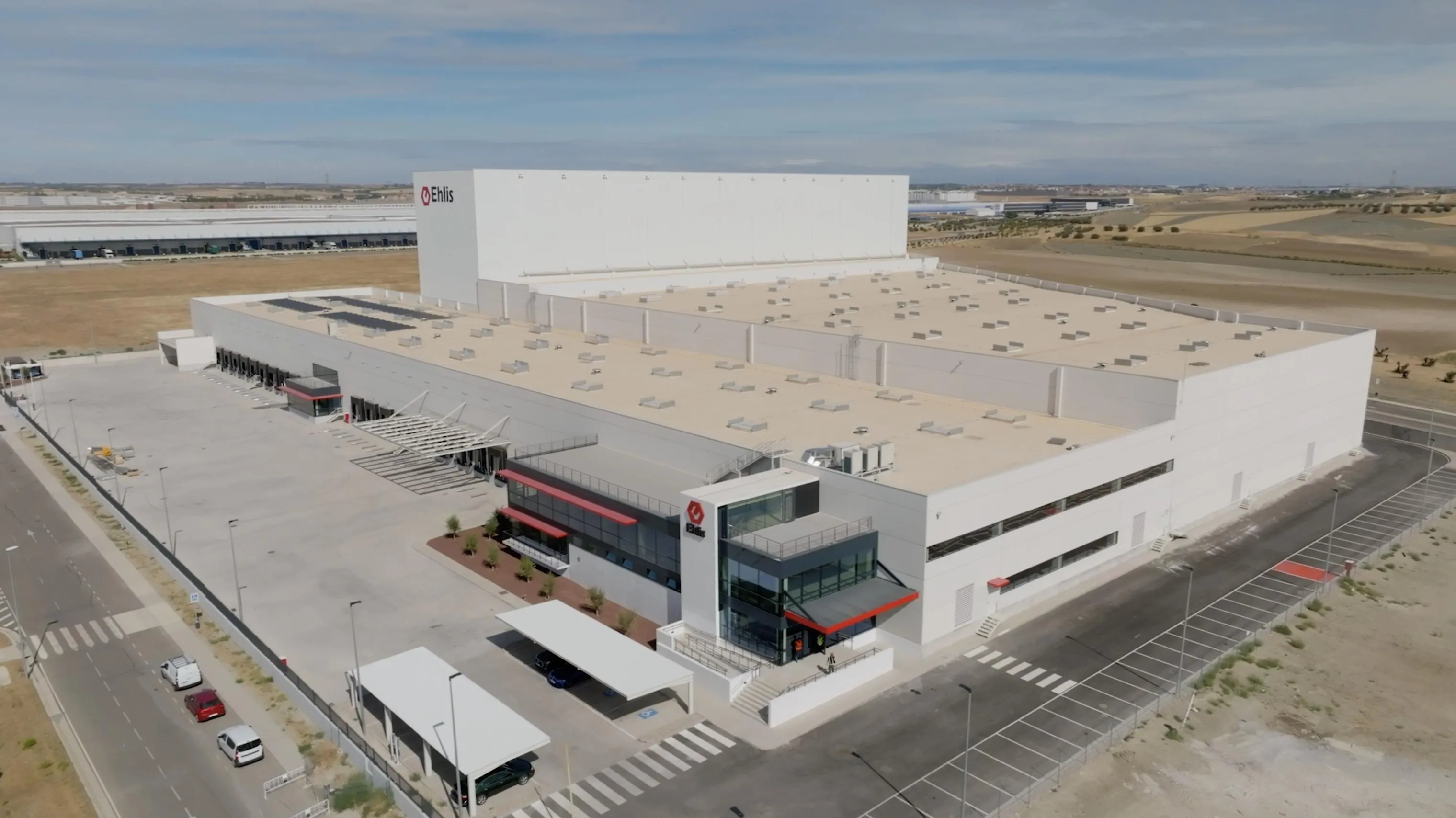 Vista aérea del centro logístico de Ehlis en Illescas: una gran nave blanca con el rótulo «Ehlis», muelles de carga, aparcamiento y un edificio de oficinas acristalado en la entrada.