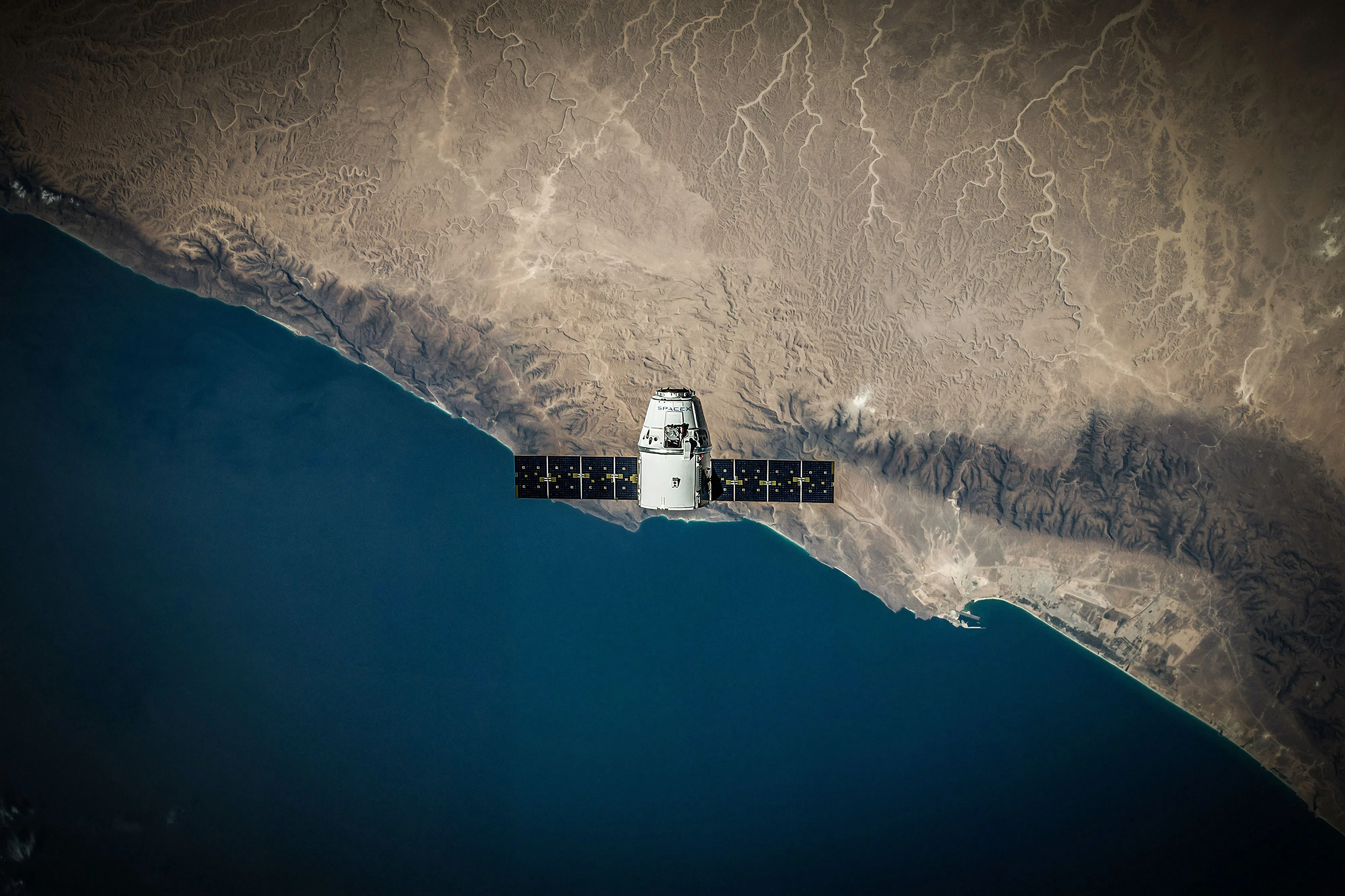 Cápsula espacial con los paneles solares desplegados, en órbita sobre una costa desértica y el mar.