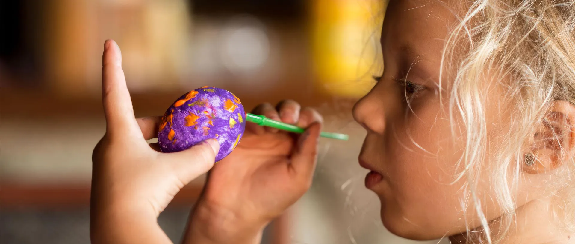 Primer plano de una niña rubia pintando un huevo de plástico morado con un pincel verde, sujetándolo con la otra mano.