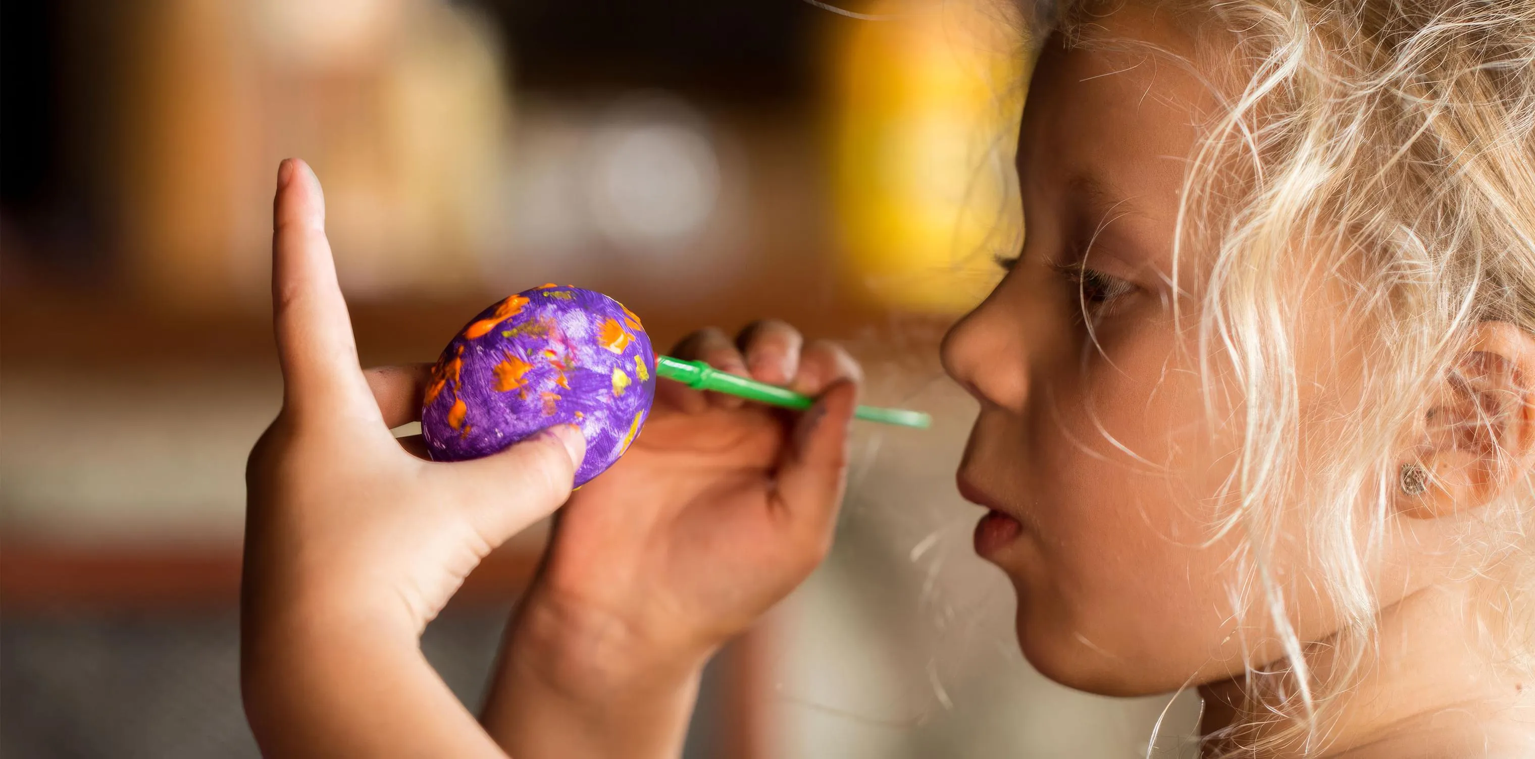 Primer plano de una niña rubia pintando un huevo de plástico morado con un pincel verde, sujetándolo con la otra mano.