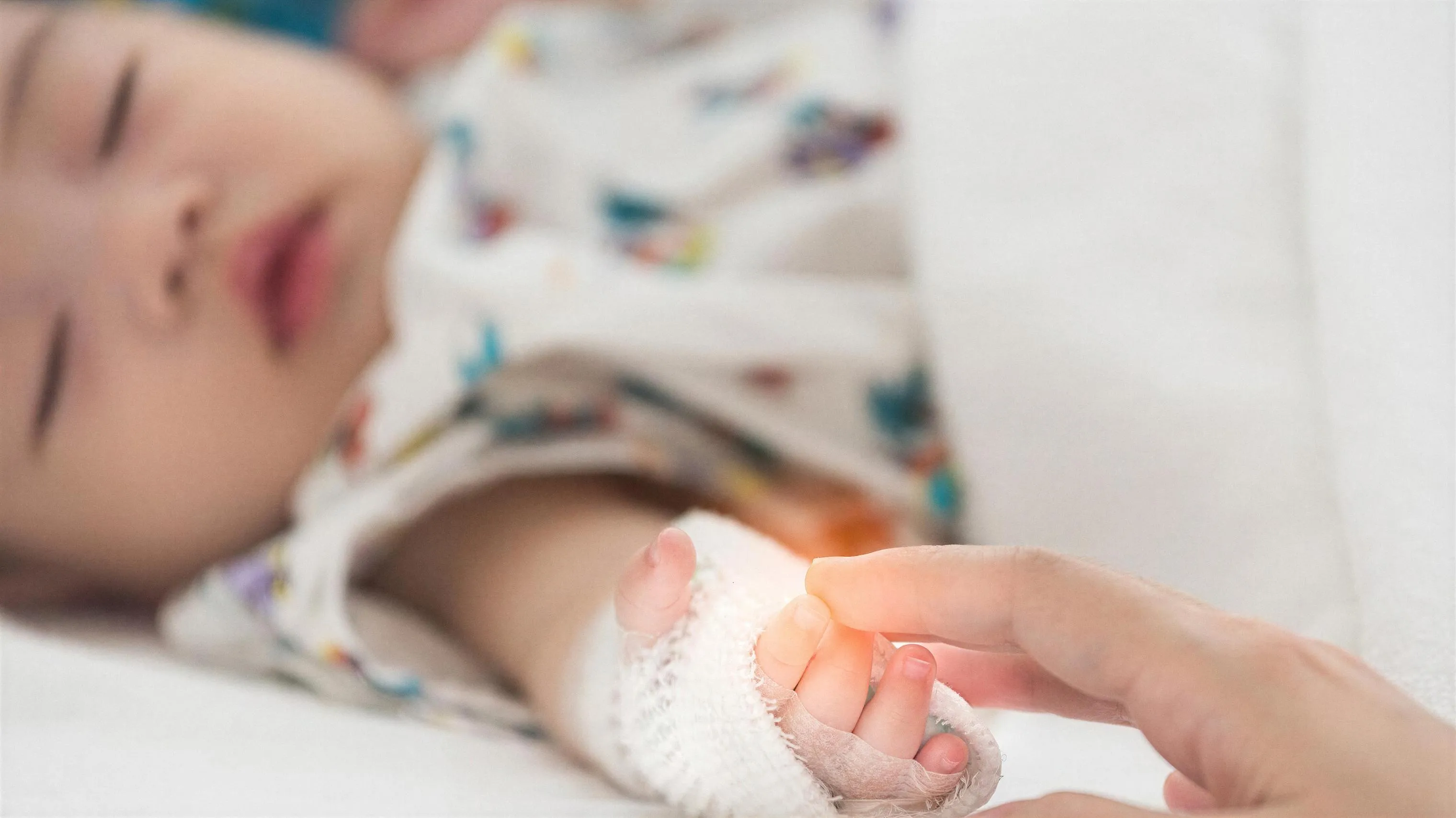 Un bebé dormido en una cama de hospital, con la mano vendada, sujeta con sus deditos el dedo de un adulto.