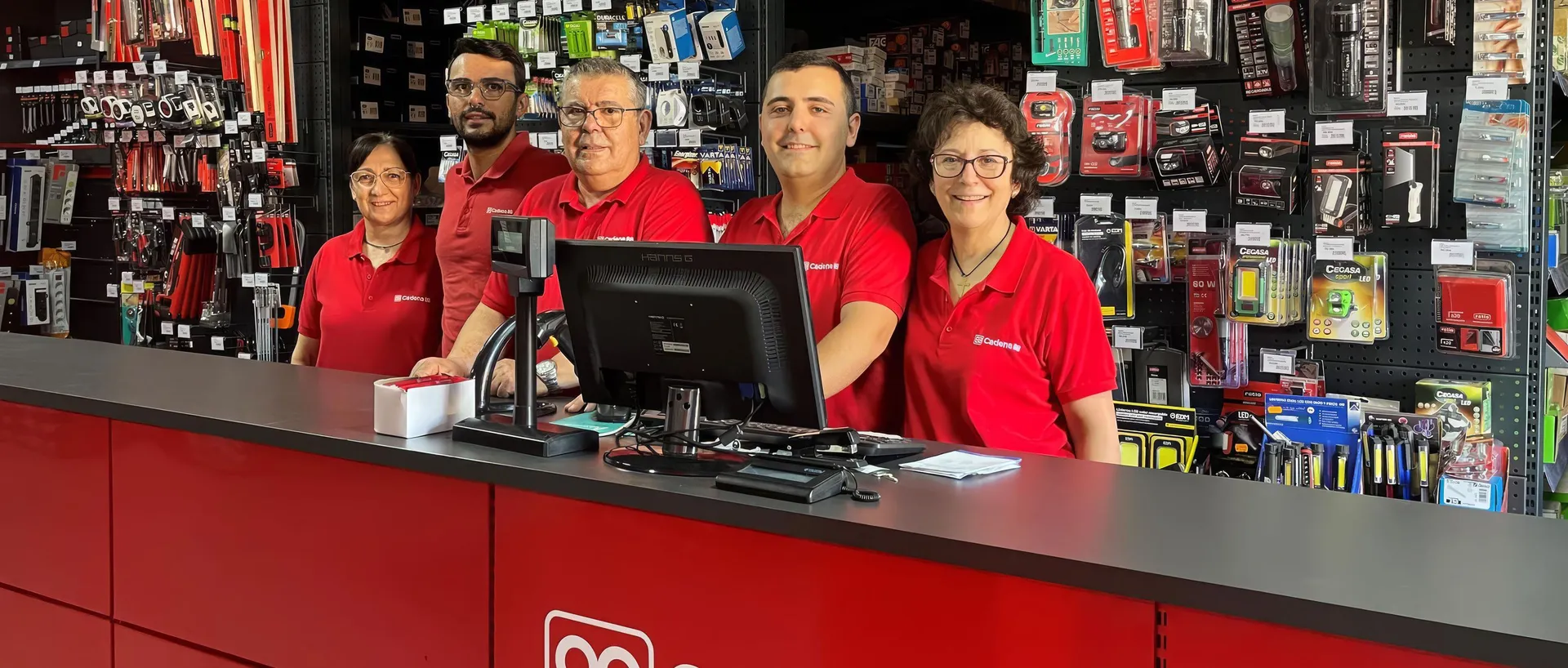 Cinco empleados de una ferretería Cadena 88, tres hombres y dos mujeres con polo rojo de la marca, sonríen detrás del mostrador, con las estanterías de herramientas y accesorios detrás.