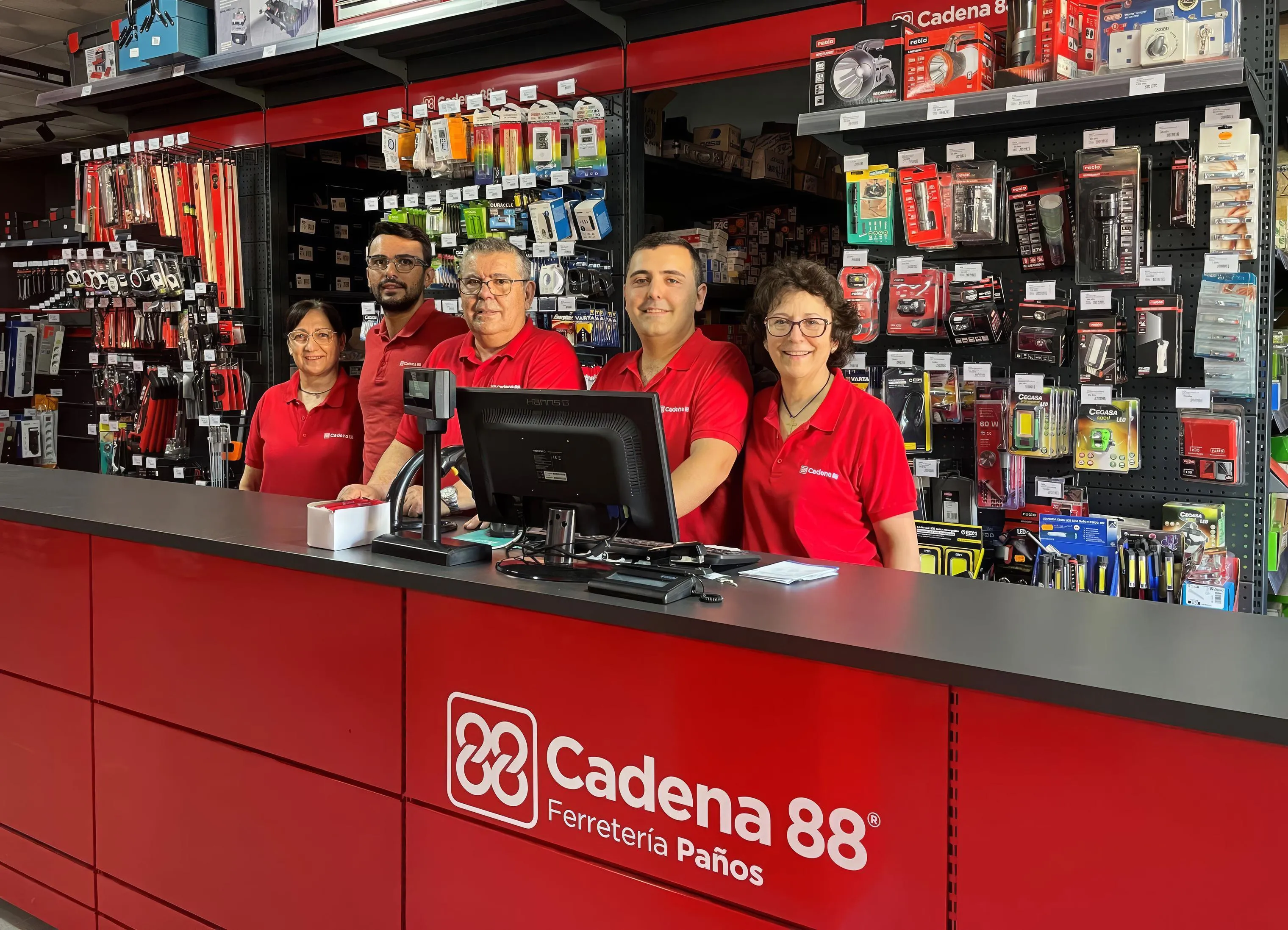 Cinco empleados de una ferretería Cadena 88, tres hombres y dos mujeres con polo rojo de la marca, sonríen detrás del mostrador, con las estanterías de herramientas y accesorios detrás.
