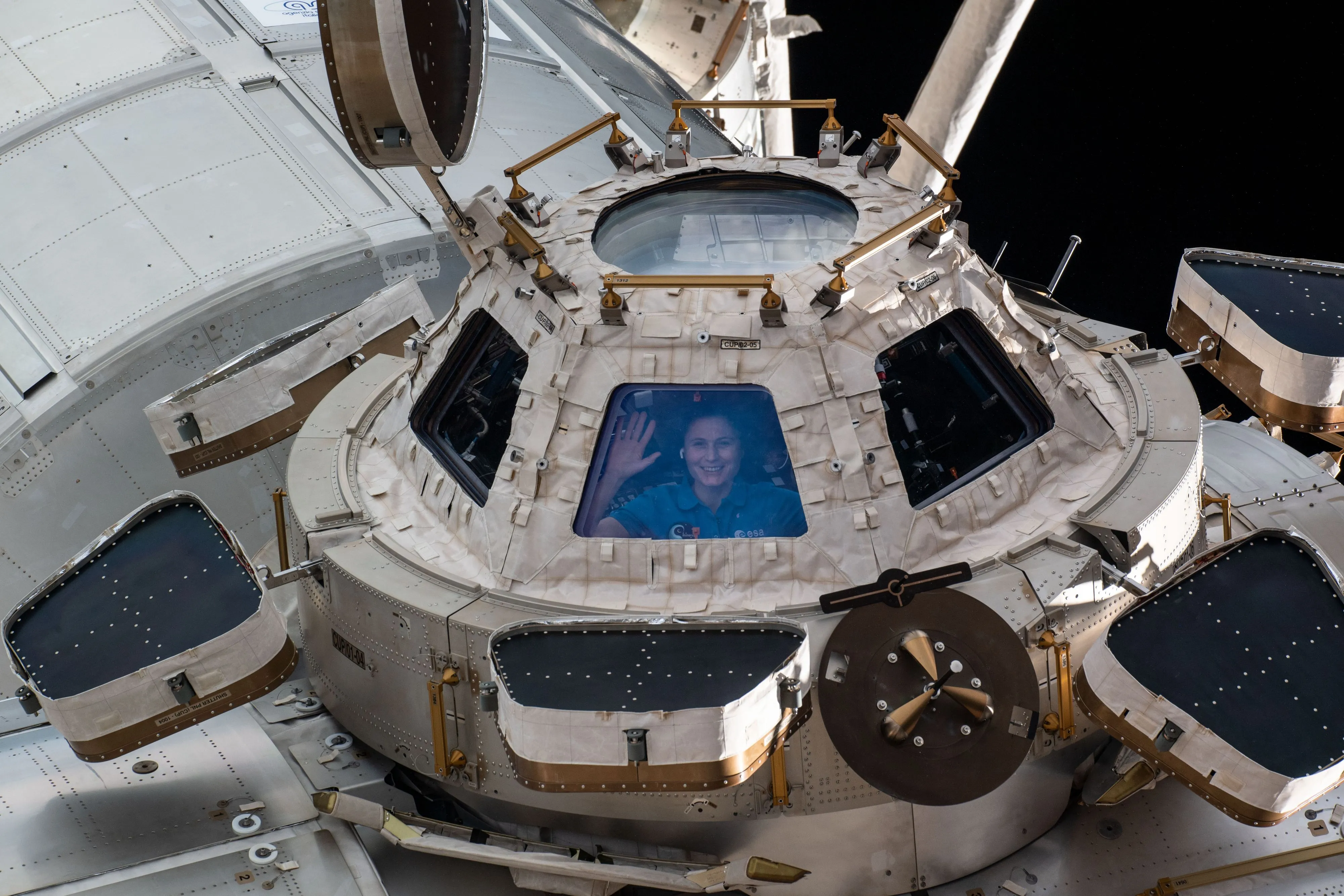 Una astronauta saluda desde una de las ventanas de la cúpula de observación de la Estación Espacial Internacional, vista desde el exterior.