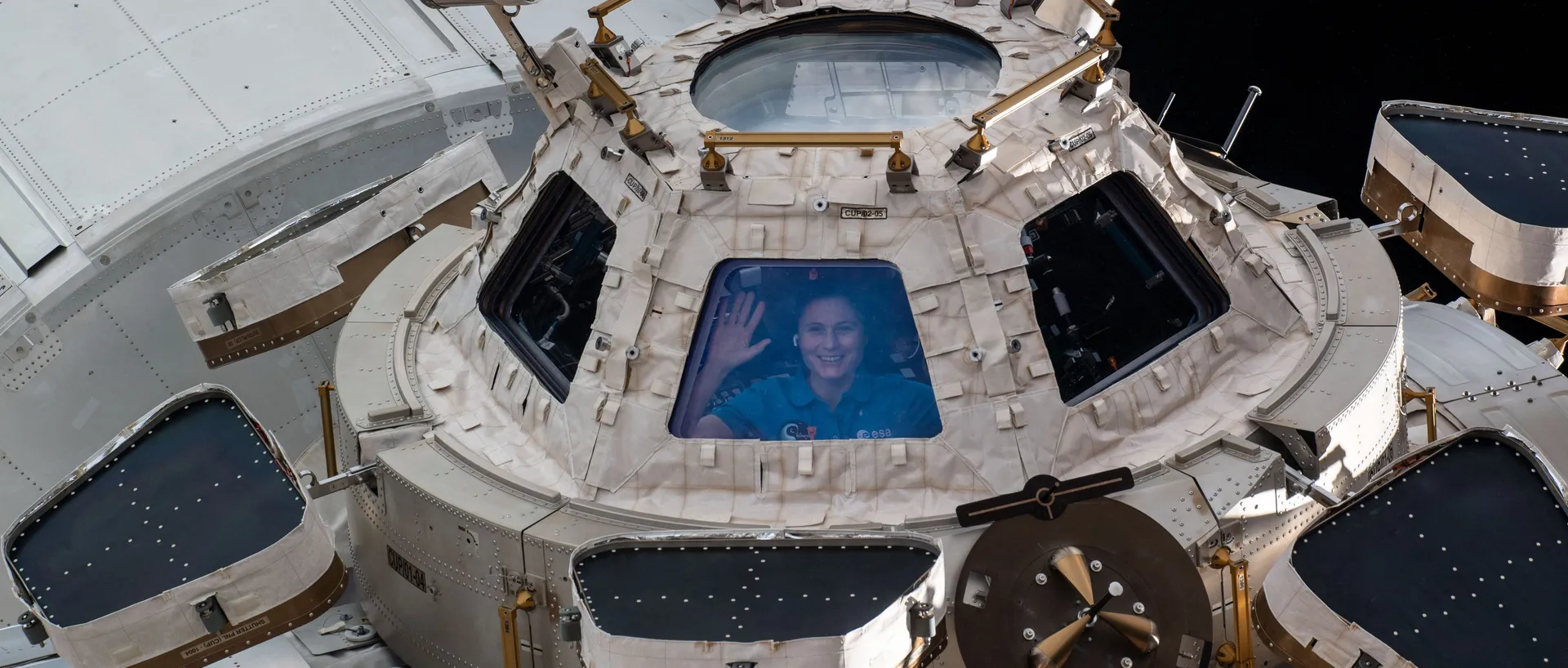 Una astronauta saluda desde una de las ventanas de la cúpula de observación de la Estación Espacial Internacional, vista desde el exterior.