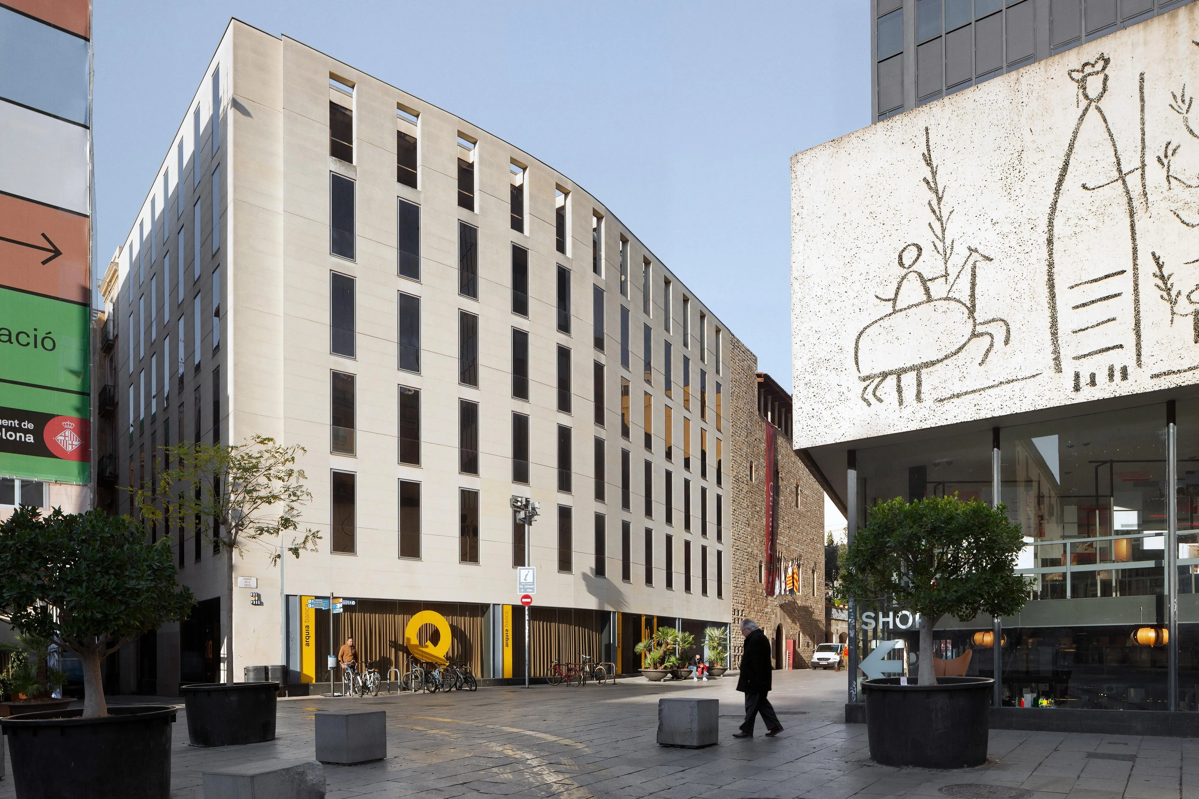 Fachada de piedra clara de la sede del Col·legi d'Arquitectes de Catalunya en la plaza Nova de Barcelona, con el friso de Picasso a la derecha y un peatón cruzando la plaza