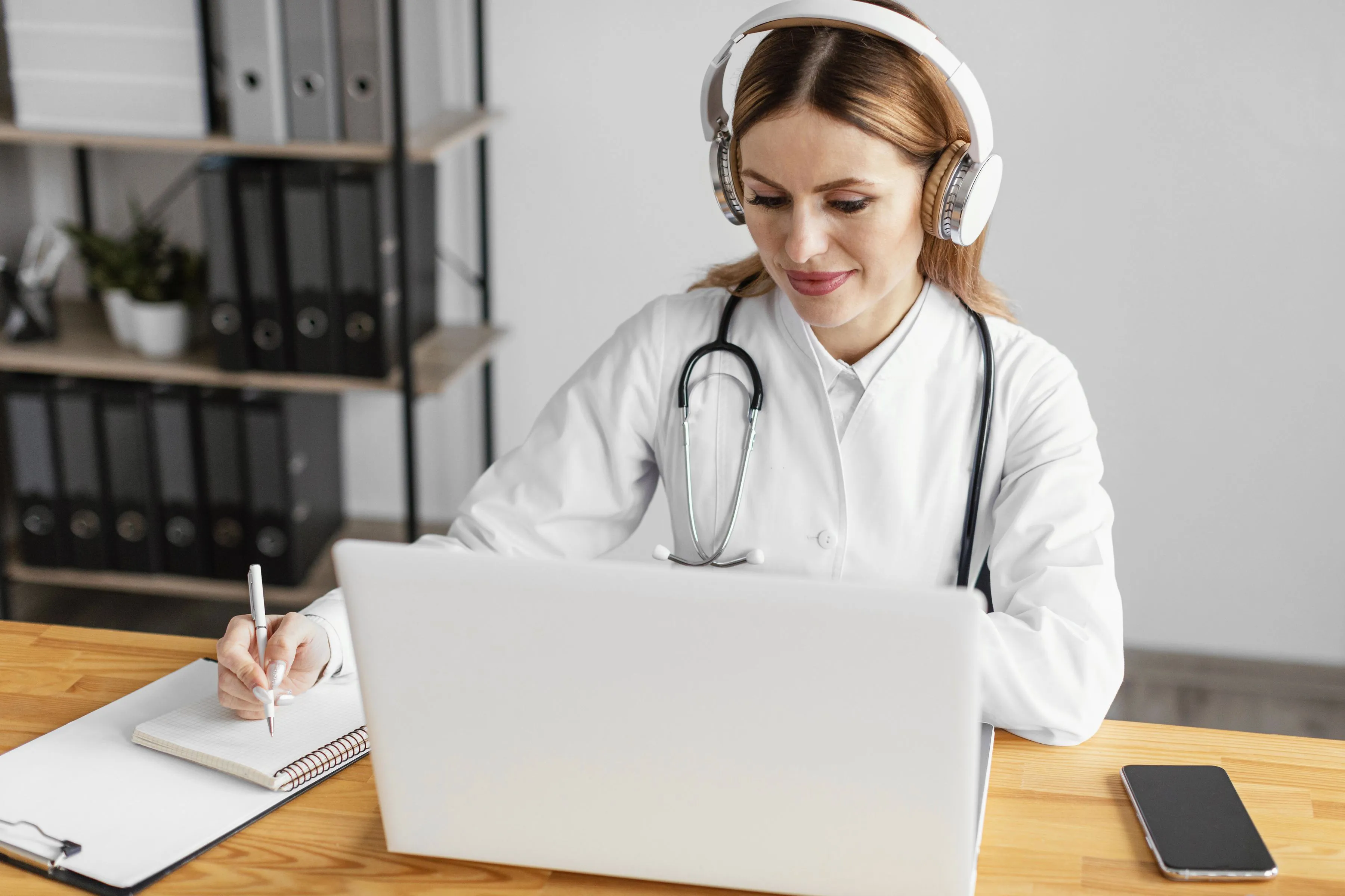 Una doctora con bata blanca, fonendoscopio y auriculares sigue algo en su portátil mientras toma apuntes en una libreta, sentada a una mesa de madera en su despacho.