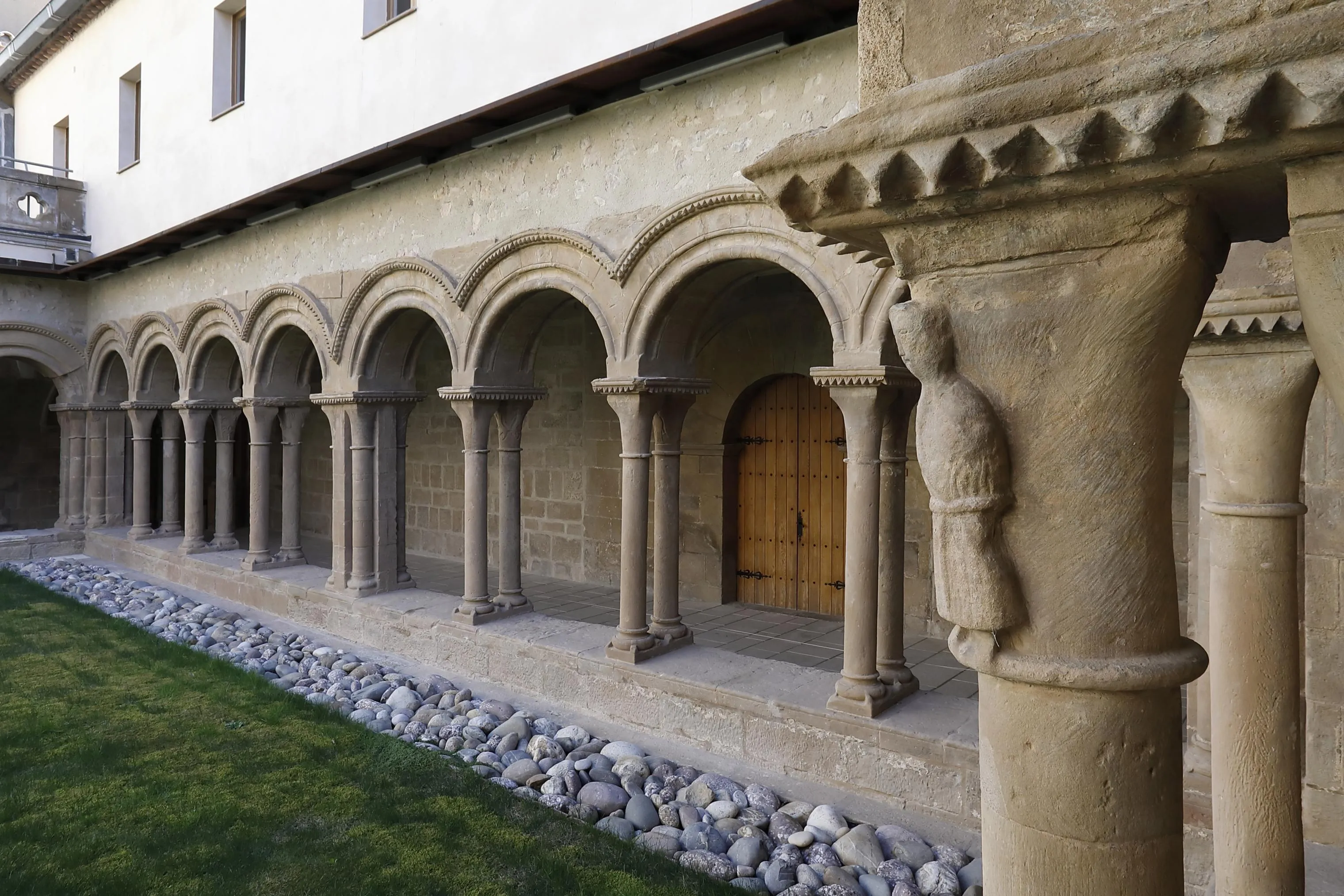 Claustro románico de piedra del Monestir de les Avellanes, con una arcada de columnas, una puerta de madera al fondo y césped con piedras de río en primer plano; no hay personas.