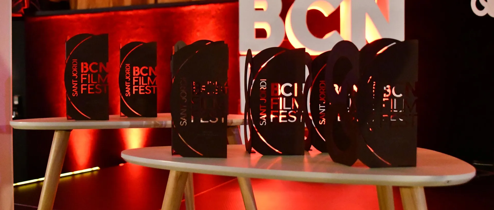 Varios trofeos negros del BCN Film Fest con el texto «Sant Jordi BCN Film Fest» en rojo, colocados sobre dos mesas auxiliares blancas de patas de madera, con las letras «BCN» iluminadas en rojo al fondo.
