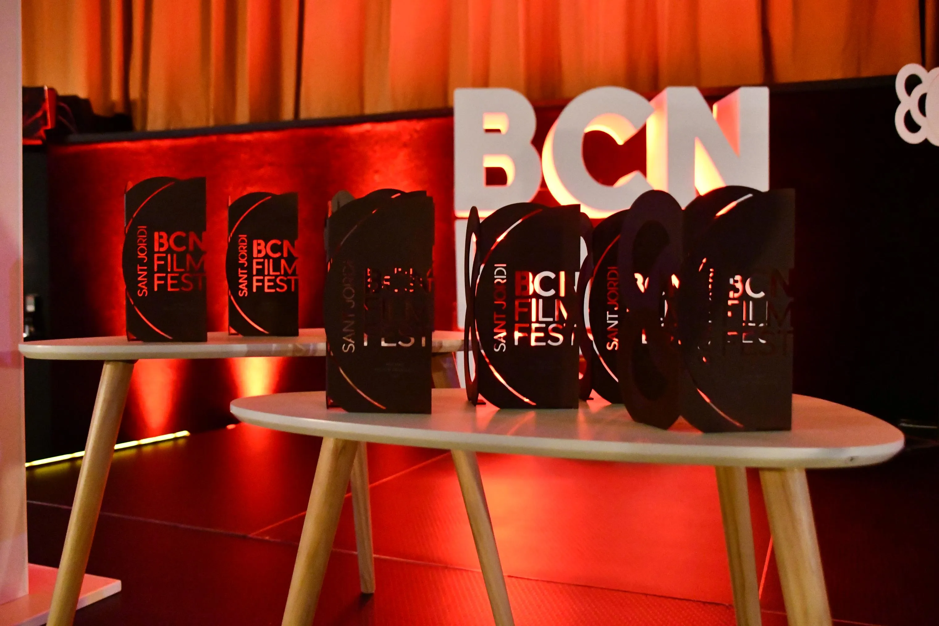 Varios trofeos negros del BCN Film Fest con el texto «Sant Jordi BCN Film Fest» en rojo, colocados sobre dos mesas auxiliares blancas de patas de madera, con las letras «BCN» iluminadas en rojo al fondo.