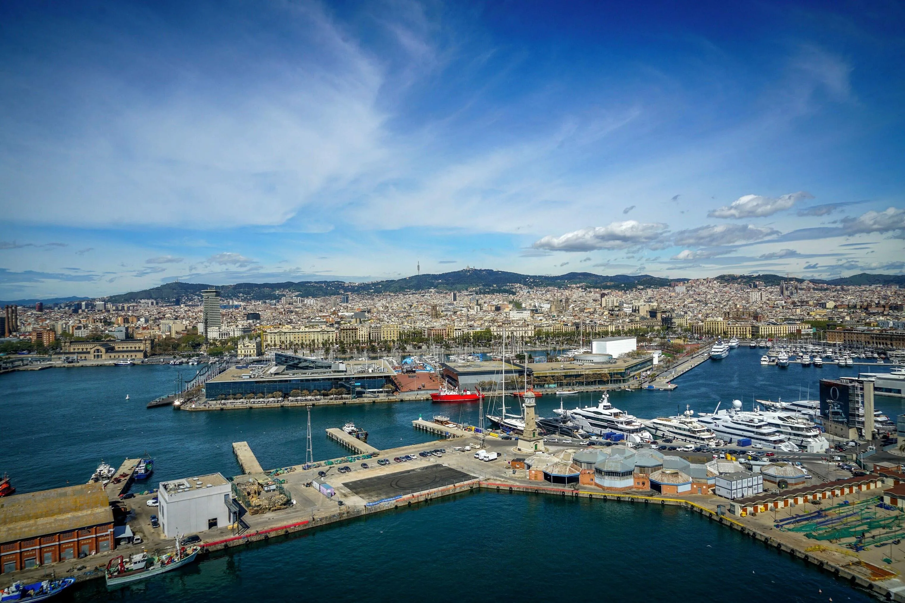 Vista aérea del Port Vell de Barcelona con los muelles deportivos llenos de embarcaciones, los edificios del Port Vell y la ciudad y las montañas de Barcelona al fondo; no se distinguen personas.