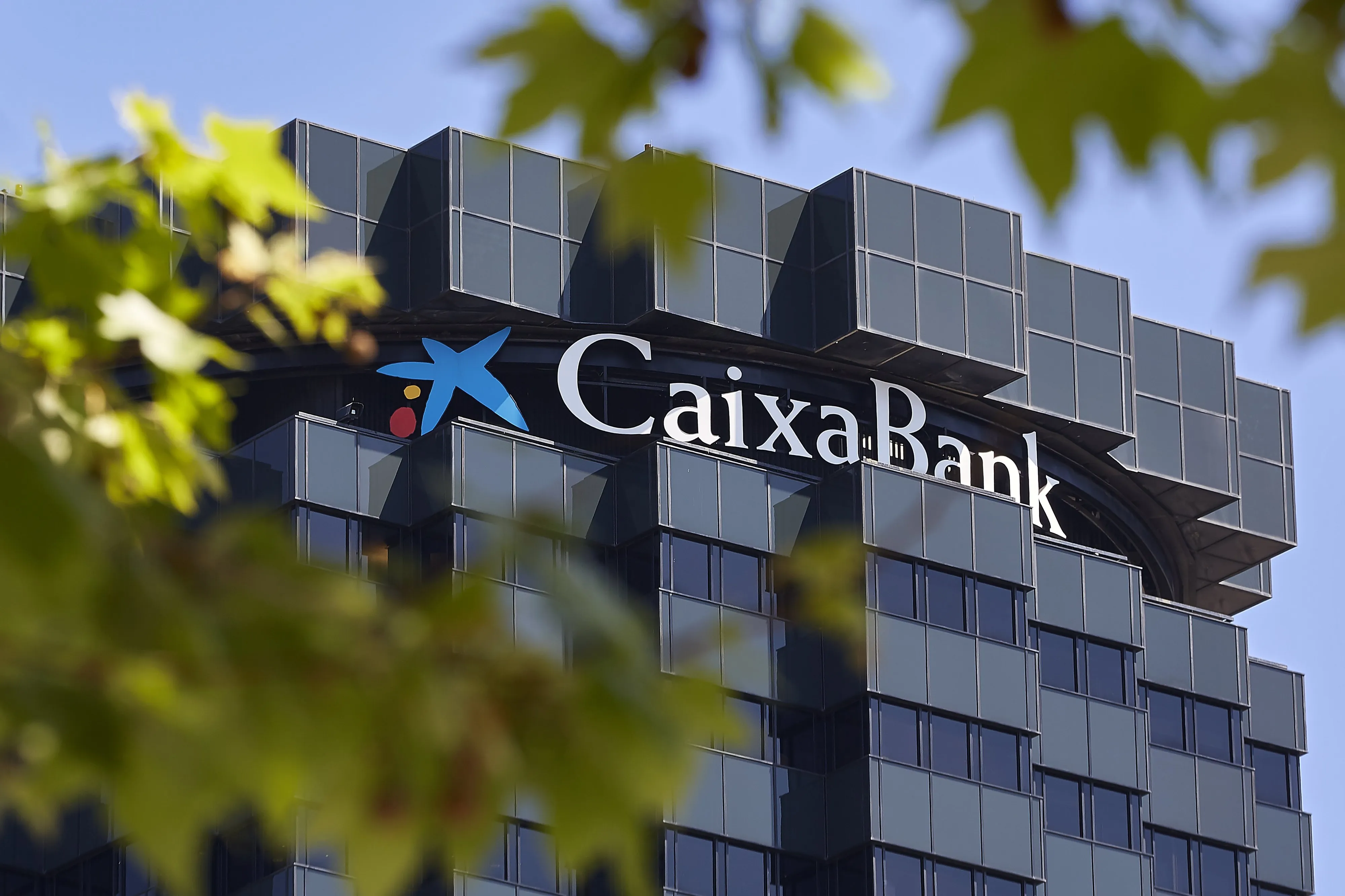 Lo alto de la torre de oficinas de CaixaBank, con el logotipo de la estrella azul y el nombre en letras blancas sobre la fachada de cristal oscuro, enmarcada por hojas de árbol desenfocadas en primer plano.