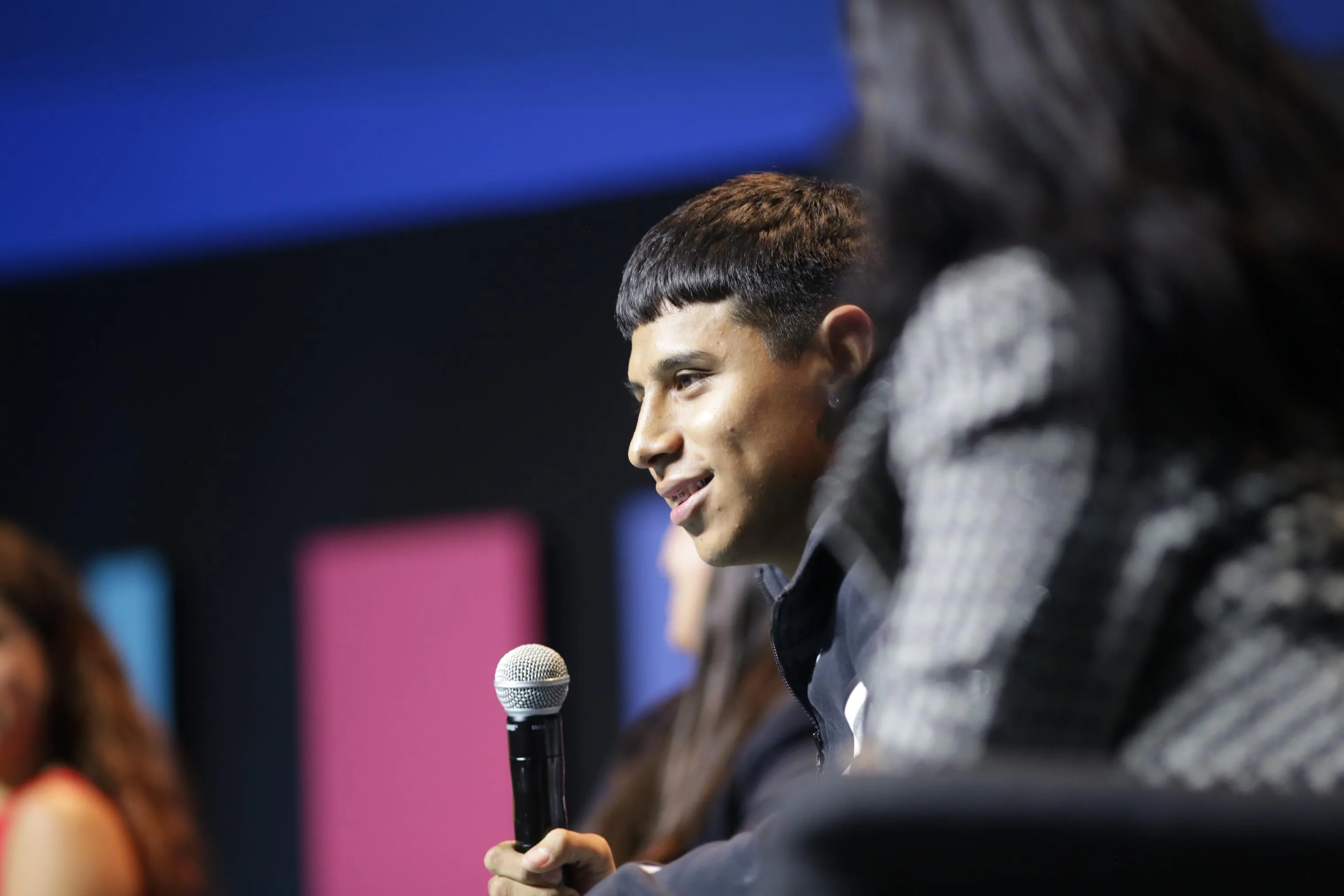 Un chico joven sonríe mientras habla por un micrófono de mano en un panel, con un fondo desenfocado de paneles azul, rosa y negro y, en primer plano borroso, la cabeza de otra persona del público.