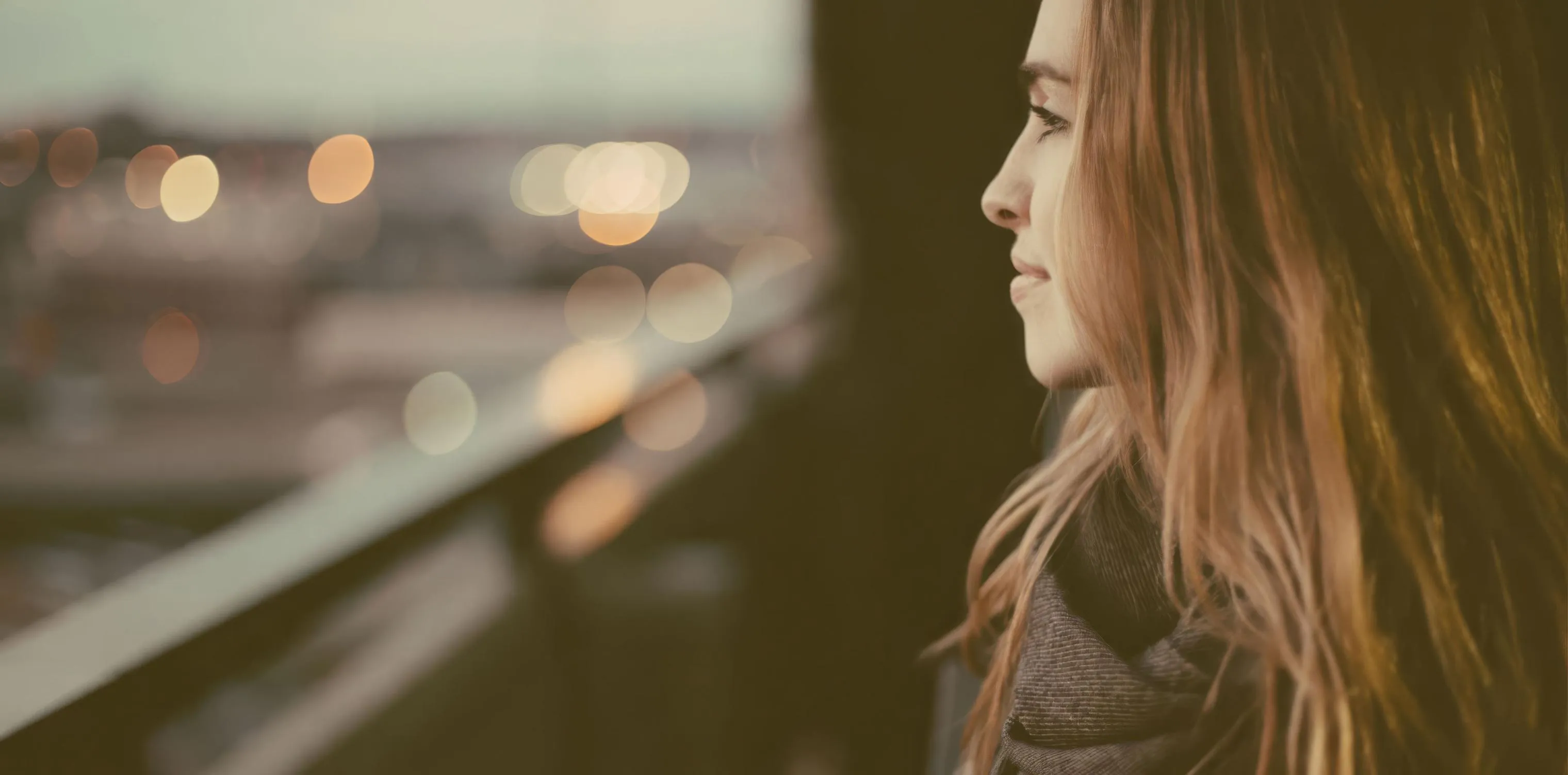 Una mujer joven, de perfil, mira hacia una ciudad al atardecer desde una terraza, con las luces de los edificios desenfocadas al fondo.