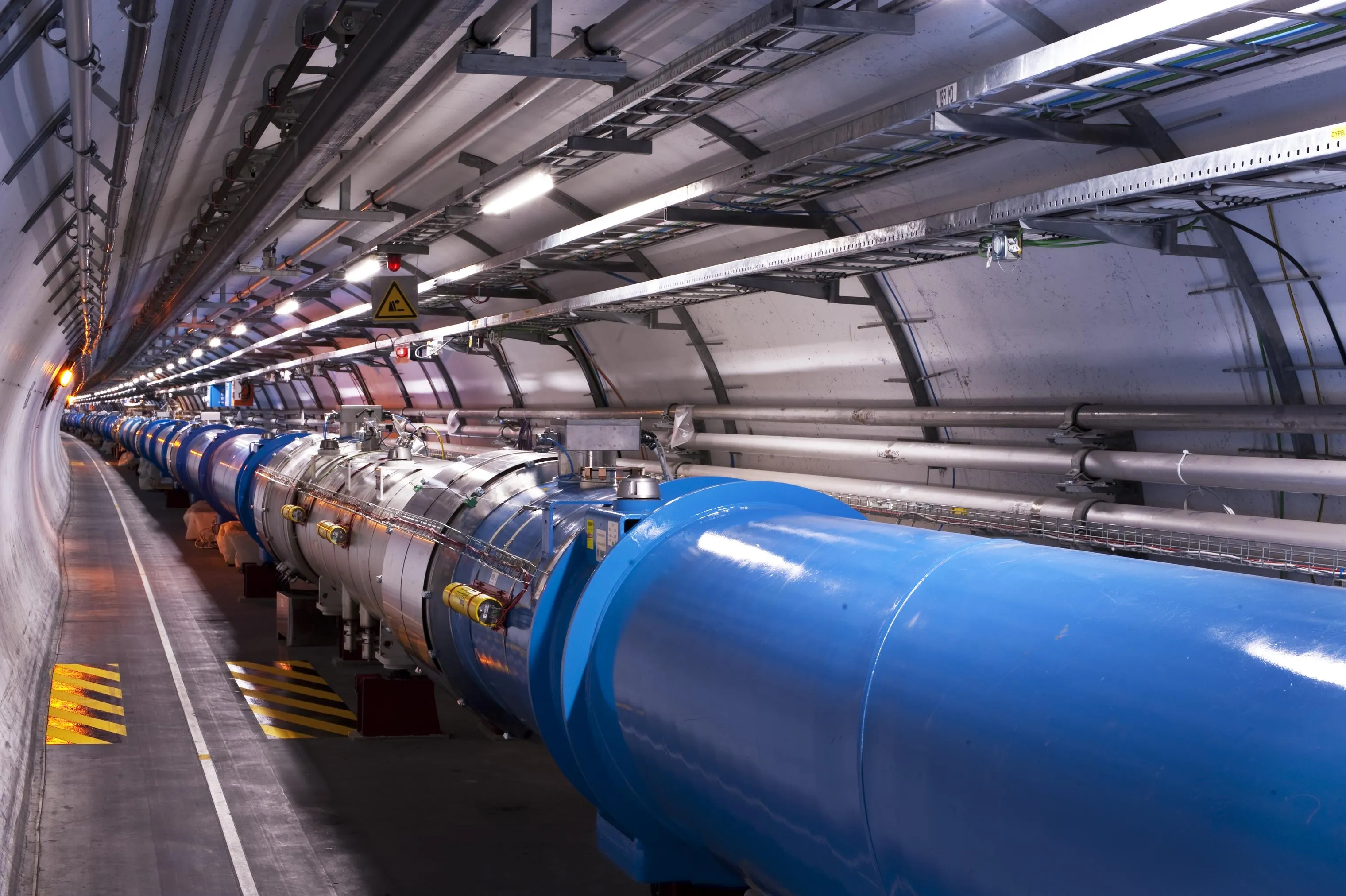 Túnel del LHC con una hilera de imanes cilíndricos azules y plateados conectados entre sí, tuberías y bandejas de cables en el techo curvado, luces encendidas, una señal amarilla de advertencia y marcas de suelo a rayas amarillas y negras.
