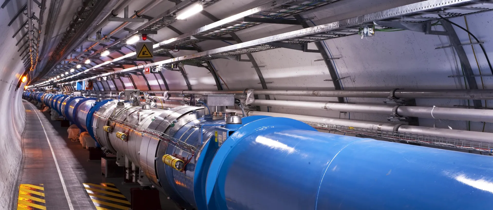 Túnel del LHC con una hilera de imanes cilíndricos azules y plateados conectados entre sí, tuberías y bandejas de cables en el techo curvado, luces encendidas, una señal amarilla de advertencia y marcas de suelo a rayas amarillas y negras.